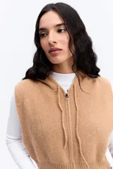 Chaleco de punto color beige, de corte corto y holgado. Presenta capucha, cierre frontal completo y terminaciones acanaladas en la cintura.