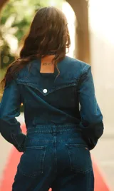 Mono de denim azul con cierre frontal de botones metálicos, cuello camisero, mangas largas y bolsillos delanteros.