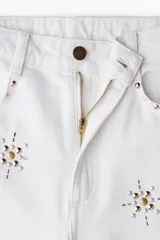 Pantalón de jean blanco, tiro superalto, con piernas amplias y corte holgado. Presenta tachas decorativas en forma de estrella o copo de nieve distribuidas por toda la prenda.