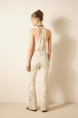 Pantalón de cuero ovino color crema, estilo jeanero con calce flare, adornado con bordados de tachas metálicas y piedras en tonos tierra y dorados a lo largo de las piernas.