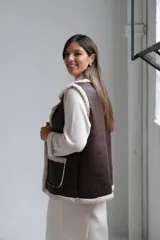 Chaleco de cuero sintético color chocolate con interior y bordes de corderito beige. Presenta solapas amplias, dos bolsillos frontales con detalle de corderito y corte recto.