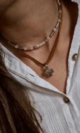 Gargantilla compuesta por un tiento de cuero marrón con dije de cruz plateada, acompañada por un collar corto de cuentas de piedra y cilindros metálicos.