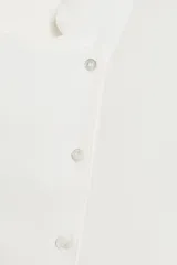 Camisa fluida de cuello redondo y manga larga con puños. Presenta un diseño con pechera frontal, cierre con botones y bajo asimétrico redondeado.