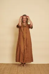 Vestido largo tipo túnica de lino color tostado, con mangas cortas abuchonadas con volados y detalles de volados verticales a lo largo de la botonera frontal oculta. Cuenta con bolsillos laterales y corte holgado.