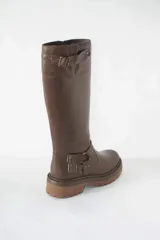 Bota de caña alta confeccionada en cuero color marrón, con suela track gruesa antideslizante. Presenta estribo decorativo con hebillas metálicas en el tobillo y detalle de ajuste con hebilla en la parte superior de la caña.