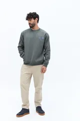 Buzo de algodón y poliéster color verde musgo, con cuello redondo y puños acanalados. Presenta un parche estampado rectangular con texto y un logo circular en el pecho.