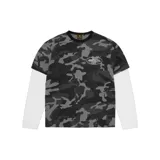 Remera de manga larga con diseño de doble capa, que combina una remera de manga corta con estampado de camuflaje en tonos grises y negros sobre una base de mangas largas blancas. Presenta un logo gráfico ovalado en el pecho.