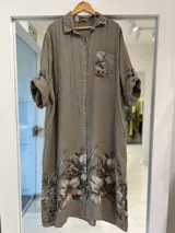 Vestido midi color visón de lino, con cierre frontal de botones, cuello camisero, mangas largas arremangables y un gran estampado floral en la parte inferior y en el bolsillo del pecho.