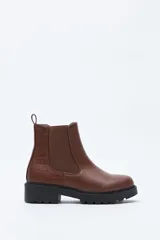 Bota tipo chelsea de caña baja con suela dentada robusta. Presenta laterales elastizados, tirador trasero y una combinación de texturas en cuero sintético liso y con relieve tipo cocodrilo en el talón.