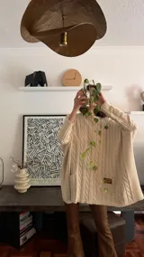 Sweater de punto color camel con diseño de trenzas verticales y acabado brillante. Presenta una silueta holgada tipo poncho, cuello redondo, mangas largas con puños ajustados y bolsillos frontales con detalle de etiqueta de cuero.