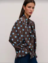 Camisa de manga larga con estampado de lunares celestes sobre fondo marrón. Presenta cuello con lazo para anudar, mangas amplias con puños acampanados y cierre frontal con botones.