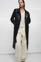 Trench de corte largo confeccionado en piel 100% con acabado brillante. Presenta cuello alto, hombreras marcadas, manga larga, bolsillos con solapa en el delantero y cierre frontal con botones. Incluye un cinturón ajustable del mismo material para entallar la silueta.