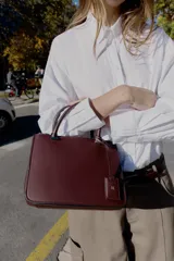 Bolso pequeño tipo satchel, color borgoña oscuro, con doble asa de mano y correa bandolera extraíble. Presenta dos compartimentos exteriores con cierre de cremallera metálica y un compartimento interior con cierre imantado.