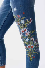 Jeans de corte ajustado y tiro medio en color azul, con detalles de bordados florales coloridos en una de las piernas.
