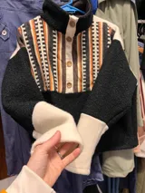 Buzo combinado con cuerpo negro de textura bouclé, mangas largas con puños anchos en tono crudo y panel frontal superior con diseño de rayas étnicas verticales en tonos crudo, negro, marrón y naranja. Cuenta con cuello alto y botonera corta frontal con botones a tono.