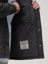 Chaqueta de hombre con diseño acolchado, confeccionada en lana Harris Tweed con patrón de tartán en tonos grises. Presenta cuello camisero, cierre frontal con botones de imitación cuero, bolsillos frontales y relleno térmico de fibras recicladas.