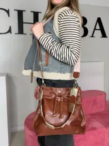 Bolso tipo bucket Chabas modelo Marin, confeccionado en cuero genuino de color marrón. Cuenta con diseño de cordón ajustable fruncido en la parte superior, manija corta decorativa de cuero con detalles metalizados y una correa larga intercambiable.