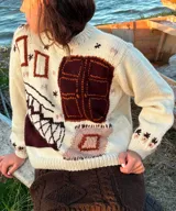 Sweater tejido en lana merino color crudo, con diseño artesanal en jacquard y bordados a mano de figuras geométricas en tonos marrón y terracota. Presenta cuello redondo, mangas largas y silueta relajada.