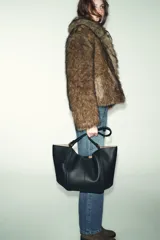Bolso tipo shopper de hombro en color beige, con diseño estructurado, doble asa y detalle de pespuntes verticales en el cuerpo. Incluye un colgante decorativo tipo etiqueta y cierre imantado.