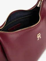 Cartera tipo hobo de Tommy Hilfiger en color bordó, confeccionada en poliuretano con acabado liso. Cuenta con compartimento principal con cierre de cremallera, bolsillo interior con cierre, asa superior y correa ajustable y extraíble. Presenta el logo metálico de la marca en relieve en la parte delantera y detalles en herrajes dorados.