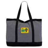 Bolso de mano tipo tote bag de lona, color gris jaspeado con base y asas negras. Presenta un parche rectangular frontal con un diseño de palmera y texto que indica "Genuine Product Of. Critical Slide. 245 Bondi Rd, Bondi, Australia".