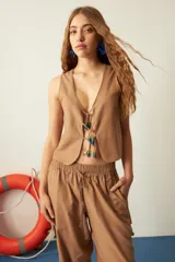 Top sin mangas color camel, con escote en V y cierre frontal mediante cordones que terminan en apliques de cuentas de vidrio azules y verdes.