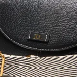 Bandolera de PU color negro con textura de cuero granulado y costuras blancas visibles. Presenta una solapa frontal con cierre metálico dorado y un logo metálico con las letras "XL". Incluye un asa superior corta y una correa larga, regulable y desmontable con estampado de cebra en blanco y negro.