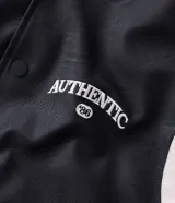 Campera bomber de cuero sintético con diseño bicolor, cuerpo negro y mangas blancas. Presenta cuello, puños y cintura con tejido elástico a rayas, cierre frontal con botones a presión y bordado con la palabra Authentic en el pecho.