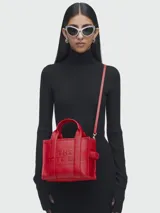 Cartera tipo tote de cuero rojo de la marca Marc Jacobs, modelo The Leather Small Tote Bag. Presenta doble asa superior, correa ajustable y extraíble para el hombro, y el texto grabado en relieve "THE TOTE BAG MARC JACOBS" en el frente.