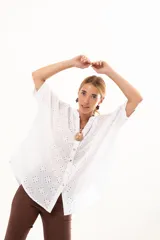 Camisa de broderie blanca con silueta oversize, escote en V, cierre frontal con botones y mangas 3/4. Presenta un diseño con bajo asimétrico, siendo ligeramente más larga en la parte posterior.