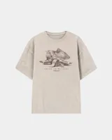 Remera de corte oversize color beige con efecto lavado, cuello redondo y mangas cortas. Presenta un estampado gráfico central en tonos oscuros con motivos de billetes y golosinas bajo la leyenda Kaboa Industries.