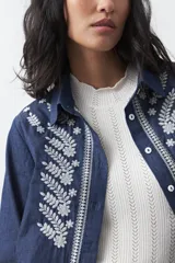 Camisa de manga larga en color azul oscuro con bordados florales en hilo blanco a lo largo de la botonadura frontal. Presenta cuello clásico y corte relajado.