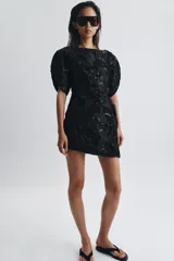 Vestido corto de cuello barco y manga corta abullonada, confeccionado en tejido negro con bordados florales y aplicaciones de abalorios y brillos a tono. Presenta un diseño con volumen en las mangas y cierre de cremallera oculta en la espalda.