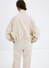Chaqueta oversize color beige con cuello alto, cierre frontal de cremallera, bolsillos frontales con solapa y mangas largas.
