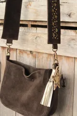 Bandolera confeccionada en cuero de gamuza marrón, con cierre superior. Presenta un llavero colgante con forma de estrella revestido en tachas metálicas y tiras flecadas en tono dorado.