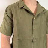 Camisa de hombre color verde oliva, de muselina con textura arrugada, manga corta, corte oversize, cuello cubano y cierre con botones. Presenta un pequeño logo bordado en el pecho.