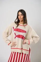 Sweater tejido color crudo con cuello redondo y mangas largas. Presenta dos peces rojos con detalles en azul bordados en el frente. Tiene ribete ondulado rojo en cuello, puños y ruedo.