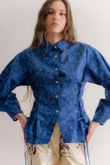 Camisa de popelina azul con estampado toile de jouy, cuello clásico y cierre frontal con botones. Presenta un diseño ajustado en la cintura mediante cordones laterales estilo corset y mangas largas abullonadas.