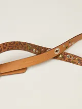Cinto de cuero con estampado animal print de leopardo, decorado con tachas metálicas, remaches y ojales. Cuenta con una hebilla cuadrada de metal y una placa metálica con el logo de la marca.
