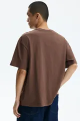 Remera marrón de algodón con cuello redondo, mangas cortas y hombros caídos, de corte holgado. Presenta un estampado frontal con la obra 'Los girasoles' de Van Gogh y el texto 'Sunflowers' en la parte superior y '-Van Gogh-' en la inferior.
