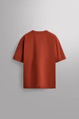 Camiseta de ajuste relajado (relaxed fit) color terracota, confeccionada en tejido técnico. Presenta cuello redondo, mangas ranglán cortas y un parche negro con logo en el bajo.