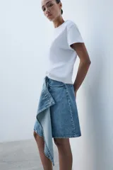 Falda de tiro medio confeccionada en denim, con trabillas, cinco bolsillos, cierre cruzado con botones y bajo acabado asimétrico.