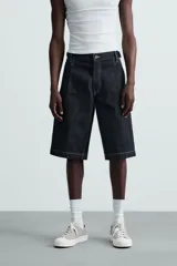 Bermuda de denim rígido color azul oscuro (sin prelavar) con corte amplio (baggy fit). Presenta pliegues frontales, trabillas ajustables en la cintura, bolsillos delanteros y bolsillos traseros de parche, todo resaltado con pespuntes a contraste.