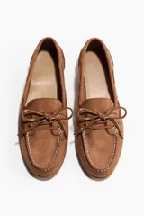 Zapatos náuticos de gamuza color marrón, con cordones frontales decorativos, costuras visibles en la puntera y ojalillos metálicos dorados. Incluyen forro interno.