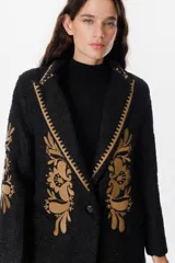 Blazer de textura suave en color crudo, con solapas clásicas y bordados florales en tonos tierra en el frente y las mangas. Presenta detalles de costura decorativa en los bordes de las solapas.