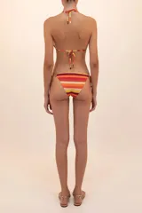 Calzón de bikini a rayas horizontales en tonos naranja, rojo, amarillo y beige. Tiene corte a la cadera, costados sin costuras, argollas doradas y tiras ajustables con detalles metálicos en las puntas.