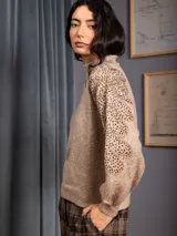 Buzo de cuello alto color beige, confeccionado en tejido de punto, con mangas largas que presentan un diseño bordado con lentejuelas en los hombros y parte superior de la manga.