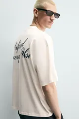 Camiseta blanca de corte relajado, con cuello redondo, manga corta y texto estampado en contraste en el pecho.