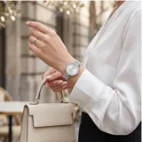 Reloj analógico de mujer con caja redonda de metal plateado y bisel decorado con cristales. Presenta una esfera minimalista con índices simples y una malla de eslabones de acero inoxidable con cierre desplegable.