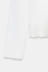 Chaqueta de punto acanalado en color blanco, con cuello redondo, manga larga y cierre frontal con botones nacarados. Presenta terminaciones con ondas en el ruedo y los puños.